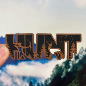 May include: A brown and black wooden cutout sign featuring the word HUNT in bold letters. The design incorporates silhouettes of a person using a bow and arrow, a deer with antlers, pine trees, and a circular crosshair target symbol.