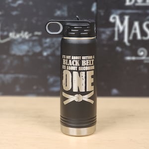 May include: Black stainless steel water bottle with a black lid and a white handle. The bottle has a white inscription that reads "It's not about getting a black belt, it's about becoming one." A white belt is tied in a knot below the text.