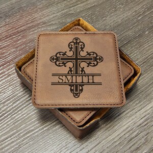May include: Set of four square brown leather coasters with a decorative cross design and the name "SMITH" engraved. The coasters are in a matching brown leather box with a yellow interior.