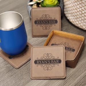 May include: Set of four brown leather coasters with a decorative design and the text "Ryan's Bar" engraved on each coaster. The coasters are in a brown leather holder.