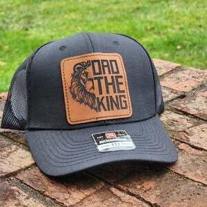 May include: A dark gray trucker hat with a black mesh back and a brown leather patch. The patch features a lion's head and the words "DAD THE KING" in a bold font. A small tag is visible on the brim.