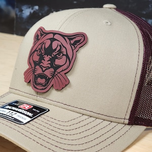 May include: A tan and burgundy trucker hat with a cougar patch. The hat features a mesh back, a curved brim, and a patch of a cougar's face. The Richardson 112 adjustable trucker hat is a classic style.
