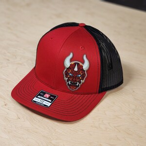 May include: Red and black trucker hat with a red and yellow embroidered Japanese demon mask on the front. The hat has a black mesh back and a red front panel. The hat is adjustable and has a Richardson 112 label.