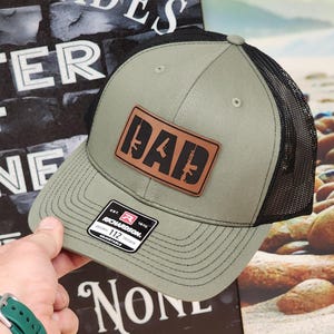 May include: A green and black trucker hat with a brown leather patch that says "DAD" with gun silhouettes. The hat has a black mesh back and a Richardson 112 adjustable strap.