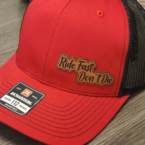 May include: Red and black trucker hat with a brown leather patch that says "Ride Fast Don't Die". The hat is adjustable and has a Richardson 112 label.
