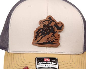 May include: A trucker hat with a tan brim, a white front panel, and a gray mesh back. The hat features a brown leather patch with a cowboy riding a horse. The patch is above a black and white label that reads "RICHARDSON 112 TRUCKER ADJUSTABLE."