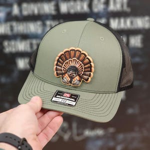 May include: A green and black trucker hat with a brown and orange turkey patch. The patch features a detailed image of a turkey with its feathers spread out. The hat has a black mesh back and an adjustable strap. The hat has a Richardson 112 label.
