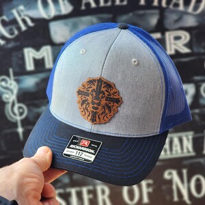 May include: A gray and blue trucker hat with a black leather patch featuring a lion with a cross and the words "Man" and "Faith". The hat is adjustable and has a black brim with blue stitching.