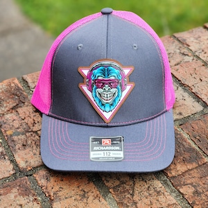 May include: A gray and pink trucker hat with a blue and white monkey wearing headphones and sunglasses graphic. The hat has a brown leather patch with the text "EST. 1970 RICHARDSON. ORIGINAL 112 TRUCKER ADJUSTABLE".