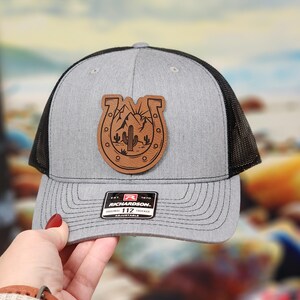 May include: Gray and black trucker hat with a leather patch featuring a horseshoe, mountains, and cacti. The hat has a curved brim and an adjustable strap. The patch is brown with a detailed desert landscape design.