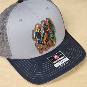 May include: A gray and black trucker hat with a patch of four horses and jockeys racing. The patch is brown with colorful accents. The hat has the text "Richardson" and "112 Adjustable" on the back.