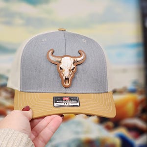 May include: A gray, tan, and white trucker hat with a bull skull patch. The hat has a curved brim and an adjustable snap closure. The patch is a detailed, brown bull skull.