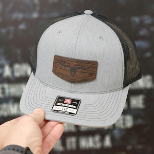 May include: Gray and black trucker hat with a brown wood-look patch featuring a skull and horns. The hat has an adjustable strap and a Richardson 112 Original Trucker label.