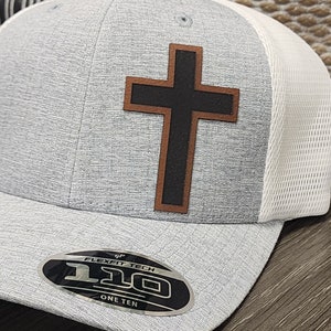 May include: A gray and white baseball cap with a black leather cross on the front. The cap has a Flexfit Tech logo and the words "One Ten" on the underside of the brim.