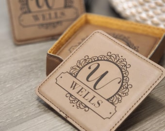 May include: A set of four brown leather coasters with a decorative monogram design. The coasters feature a cursive "W" inside a circle with a floral design and the name "WELLS" below.