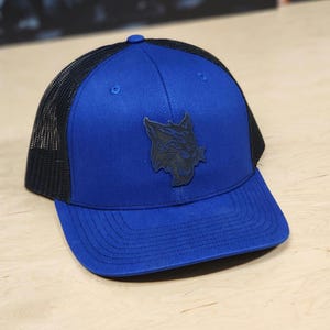 May include: A blue and black trucker hat with a black embroidered wildcat logo. The hat has a mesh back and a curved brim.