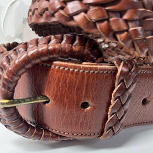 K&ouml;nnte beinhalten: Ein brauner Lederg&uuml;rtel mit geflochtenem Riemendesign und einer passenden, mit geflochtenem Leder &uuml;berzogenen Schnalle. Der G&uuml;rtel verf&uuml;gt &uuml;ber einen Metalldorn und einen glatten Lederabschnitt mit einem Einstellloch sowie sichtbare N&auml;hte.