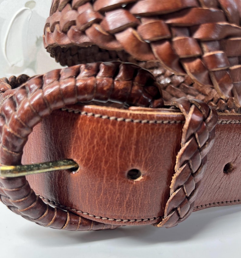 K&ouml;nnte beinhalten: Ein brauner Lederg&uuml;rtel mit geflochtenem Riemendesign und einer passenden, mit geflochtenem Leder &uuml;berzogenen Schnalle. Der G&uuml;rtel verf&uuml;gt &uuml;ber einen Metalldorn und einen glatten Lederabschnitt mit einem Einstellloch sowie sichtbare N&auml;hte.