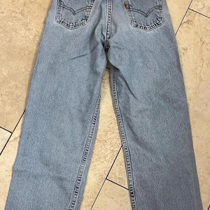 May include: Light blue denim jeans with classic five-pocket styling. The jeans feature a faded wash, back pockets with signature stitching, and a brown leather patch. The jeans are laid flat, showcasing the back view.