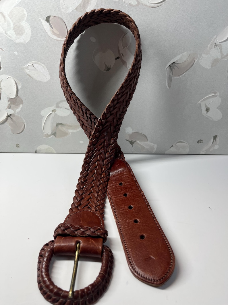 K&ouml;nnte beinhalten: Ein dunkelbrauner geflochtener Lederg&uuml;rtel mit einer runden, mit Geflecht &uuml;berzogenen Schnalle und einem glatten, festen Lederende mit f&uuml;nf gestanzten Einstelll&ouml;chern. Der G&uuml;rtelriemen besteht aus mehreren ineinander verschlungenen Lederstr&auml;ngen.