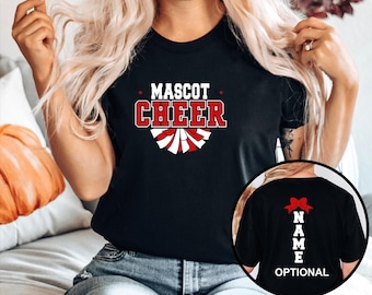 May include: A black t-shirt featuring the white and red text MASCOT CHEER above a graphic of a pom-pom. An inset circle shows the back of the shirt with a red bow graphic above the vertical white text NAME and the word OPTIONAL below it.
