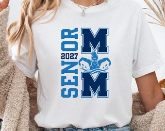 Peut inclure: Un t-shirt blanc à manches courtes avec un motif graphique bleu. Le texte indique SENIOR 2027 avec une grande lettre M au-dessus et au-dessous d'une icône de nageur. Le nageur est représenté en position de papillon dans les éléments graphiques.