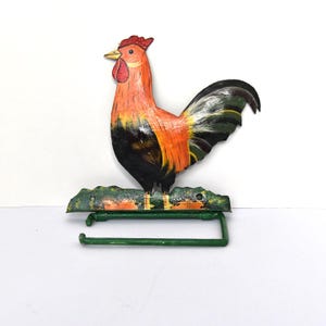 May include: A colorful rooster-shaped toilet paper holder. The wooden rooster is painted with orange, black, yellow, and green hues. The holder has a green metal bar for the toilet paper roll and a green base.