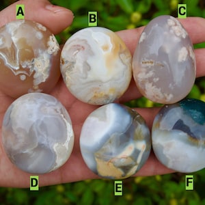 May include: A hand holds six polished, oval-shaped stones. The stones display a variety of colors, including beige, white, gray, and green, with unique patterns and swirls. The stones are smooth and appear to be gemstones or decorative objects.
