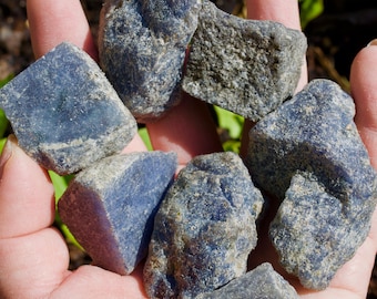 May include: A handful of rough, natural blue kyanite stones. The stones vary in size and shape, with a mix of deep blue and gray hues. The kyanite has a textured, crystalline appearance, and is held in an open hand.