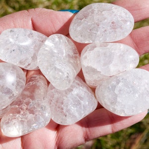 May include: Seven smooth, white, tumbled quartz stones with a cloudy, translucent appearance. The stones are in a hand, and the background is green grass.