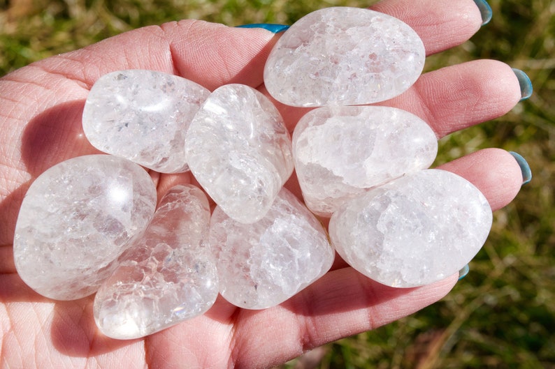 May include: Seven smooth, white, tumbled quartz stones with a cloudy, translucent appearance. The stones are in a hand, and the background is green grass.