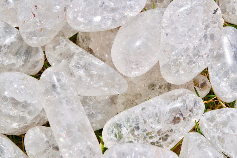 May include: A collection of clear quartz crystal stones, each with a unique shape and texture. The stones are smooth and polished, with a slight iridescent sheen.