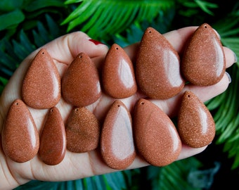 May include: A collection of eleven teardrop-shaped goldstone pendants, each featuring a smooth, polished surface with embedded metallic sparkles. These brown, glittery gemstone charms are arranged in two rows, each with a small hole at the top.