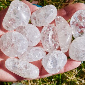 May include: A collection of ten clear quartz tumbled stones with a cloudy, milky appearance. The stones are smooth and polished, and they are arranged in a hand.