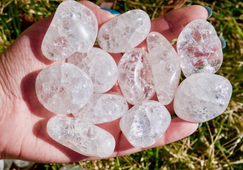 May include: A collection of ten clear quartz tumbled stones with a cloudy, milky appearance. The stones are smooth and polished, and they are arranged in a hand.
