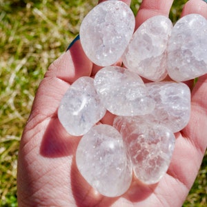 May include: A hand holding a collection of clear quartz tumbled stones. The stones are smooth and have a cloudy, milky appearance.