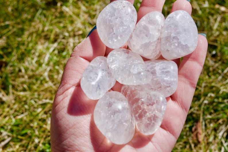 May include: A hand holding a collection of clear quartz tumbled stones. The stones are smooth and have a cloudy, milky appearance.