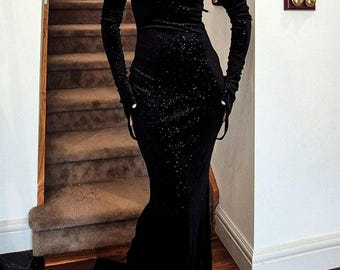 May include: A black velvet gown with a sparkling texture and long sleeves. The gown has a plunging neckline and a fitted silhouette.
