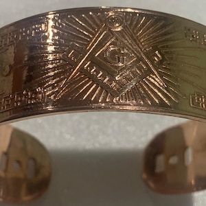 Puede incluir: Una pulsera de metal dorado con un símbolo masónico grabado en la parte delantera. El símbolo es una brújula y una escuadra con una 'G' en el centro. La pulsera tiene un diseño texturizado alrededor de los bordes.