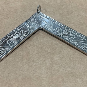 Könnte beinhalten: Silberfarbener Metall-Zirkel- und Winkel-Anhänger mit floralen und linearen Gravuren. Der Anhänger hat oben eine kleine Schlaufe zum Aufhängen. Das Design ist ein Symbol der Freimaurerei.