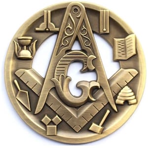 Op de afbeelding: Een bronzen vrijmetselaars-embleem met een passer, winkelhaak en de letter "G" in het midden. Het cirkelvormige ontwerp bevat een troffel, waterpas, open boek en andere symbolische gereedschappen. Het embleem heeft een gedetailleerde, antieke afwerking.