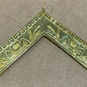 Könnte beinhalten: Eine goldfarbene, rechtwinklige Rahmenecke mit floralen und geometrischen Prägungen. Der Rahmen hat oben eine kleine Schlaufe zum Aufhängen. Das Design weist eine Kombination aus matten und glänzenden Oberflächen auf und erzeugt eine Vintage-Ästhetik.