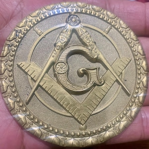 Può includere: Una moneta massonica color oro con il simbolo della Squadra e del Compasso, un emblema prominente della Massoneria. La moneta ha un design dettagliato con un bordo decorativo e dettagli intricati all'interno dei simboli centrali.