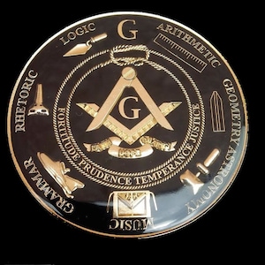 Puede incluir: Un emblema circular negro y dorado con una brújula y un cuadrado en el centro. La brújula y el cuadrado están rodeados por un diseño de cuerda y las palabras "Fortaleza Prudencia Templanza Justicia" en un círculo. Las palabras "Retórica", "Lógica", "Aritmética", "Geometría", "Astronomía", "Gramática" y "Música" están inscritas alrededor del borde exterior del emblema.