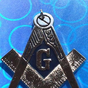 May include: A silver-colored Masonic symbol with a blue background and the letter 'G' in the center. The symbol is a square and compass with a level on top.