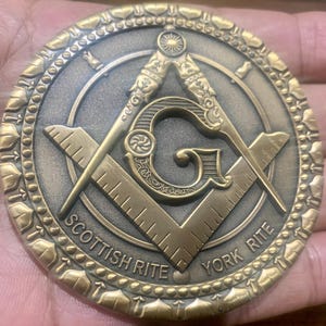 Op de afbeelding: Een bronzen vrijmetselaarsmunt met het symbool van de passer en de winkelhaak, en de woorden "Scottish Rite" en "York Rite". De munt heeft een gedetailleerde rand en een centraal ontwerp met een "G" in het midden.