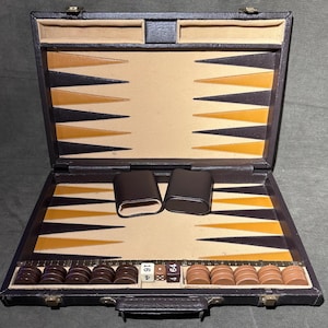 May include: Open backgammon set in a black leather case. The playing surface features alternating black, tan, and gold triangles. Includes brown and tan game pieces, two dice cups, and dice. The set is open, revealing the interior game board and accessories.