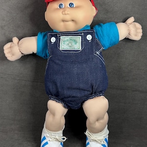 Könnte beinhalten: Eine Cabbage Patch Kids-Puppe mit roter Kappe, blauem Hemd und Jeans-Latzhose. Die Puppe hat blaue Augen und weiße Turnschuhe mit blauen Streifen. Die Latzhose der Puppe hat einen Cabbage Patch Kids-Aufnäher.