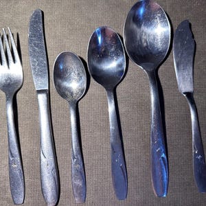 Puede incluir: Un juego de seis cubiertos de acero inoxidable, que incluye un tenedor, dos cuchillos y tres cucharas. Cada mango tiene un diseño de estrella. Los utensilios están dispuestos sobre una superficie marrón.