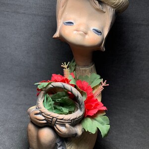 Może przedstawiać: Vintage ceramiczna figurka siedzącego dziecka w kapeluszu, trzymającego kosz kwiatów. Figurka jest pomalowana w stonowanych odcieniach z czerwonymi i zielonymi akcentami kwiatowymi. Oczy dziecka są zamknięte, a ogólna estetyka jest nostalgiczna.
